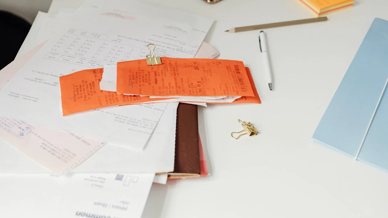 白い机の上でクリップに留められたレシートと請求書。そばにペンと水色のフォルダーがある