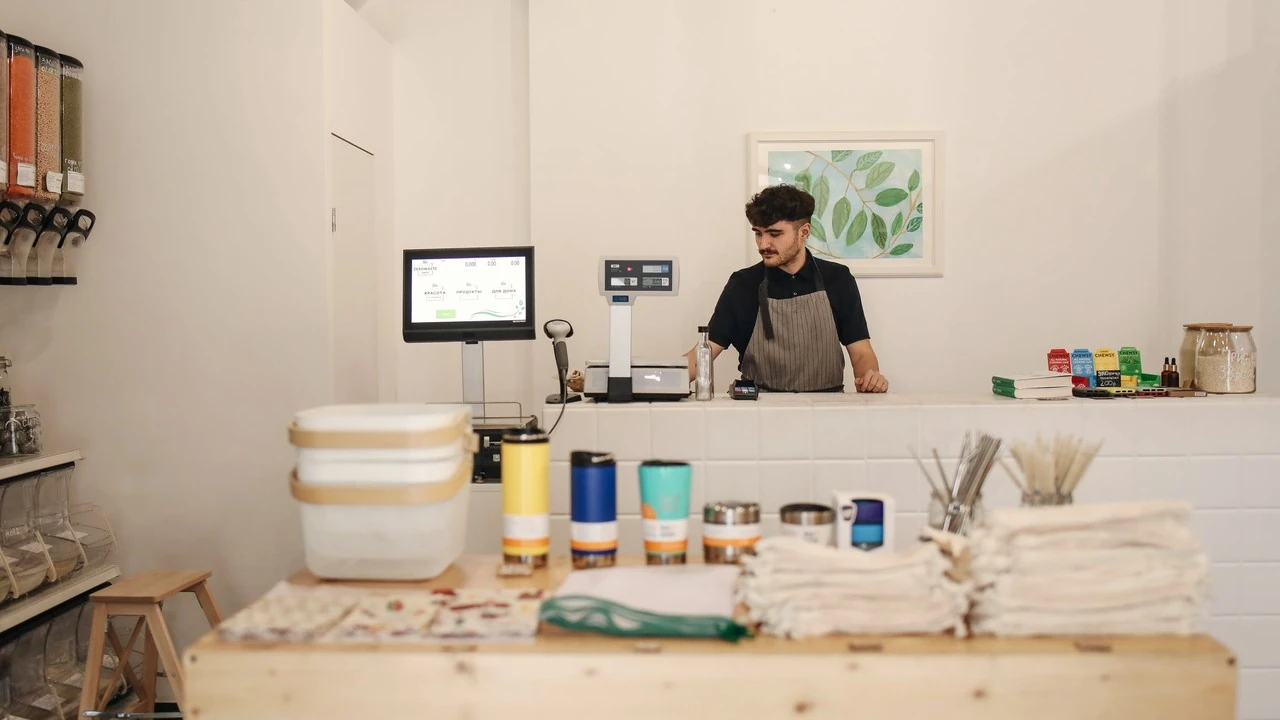 A young man at the counter of a small shop, with a till, a scale and shelves of goods around him