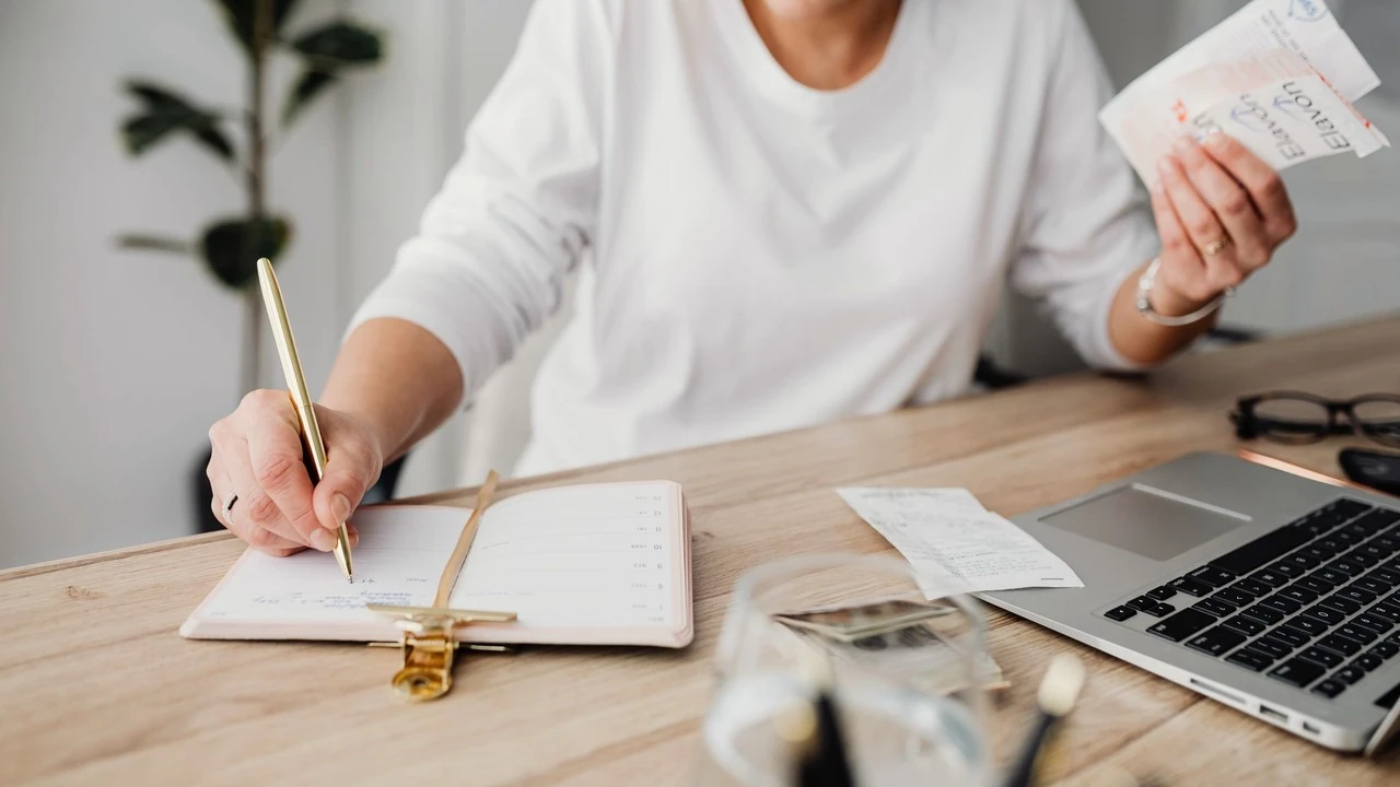 A woman writes in a notebook at her desk, a receipt in her other hand and a laptop beside her