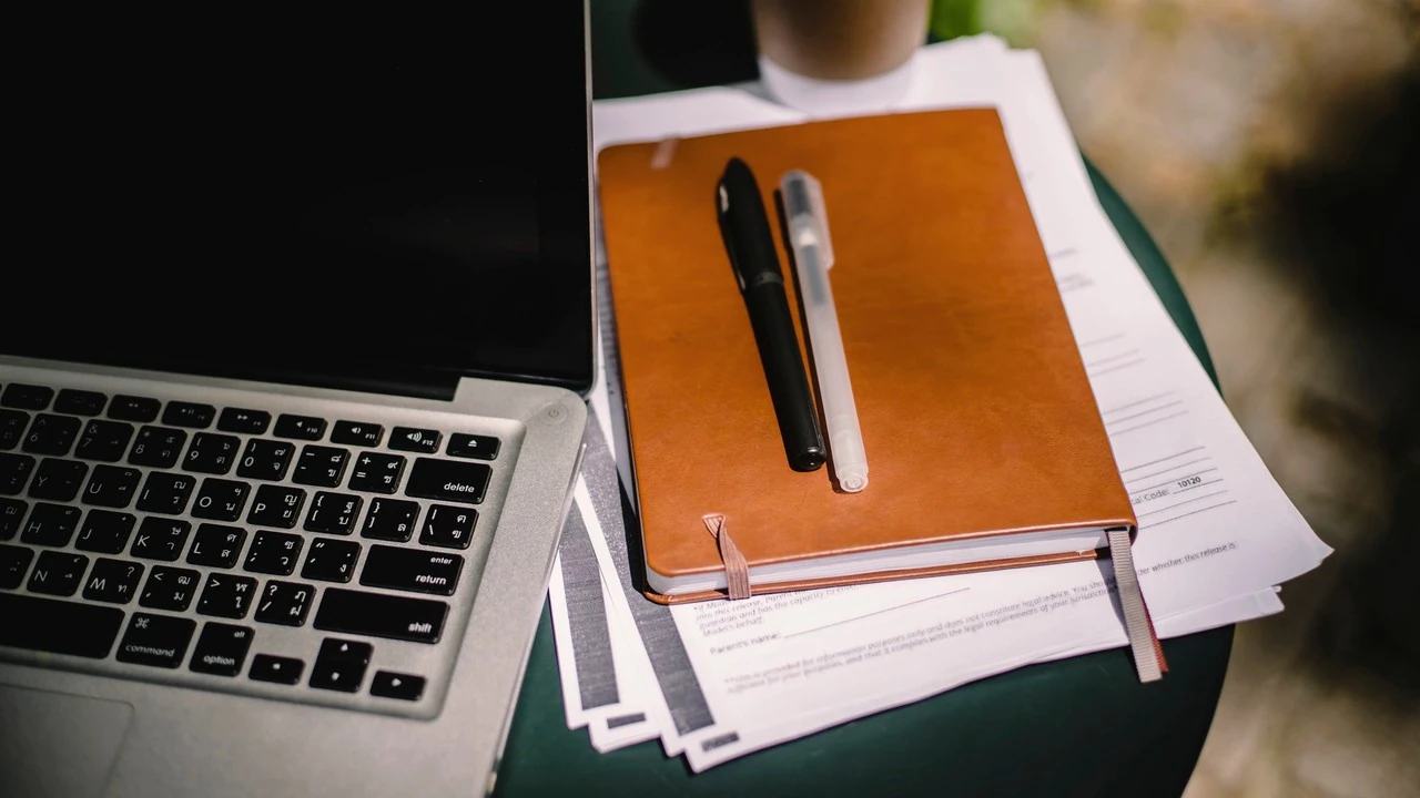 Un carnet en cuir et des stylos posés sur des feuilles volantes, à côté d'un ordinateur portable ouvert et d'une tasse de café